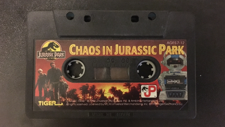 Photo de la cassette de Chaos In Jurassic Park, avec dessus une photo du 2-XL et une image des acteurs sur fond de jungle et et soleil couchant.